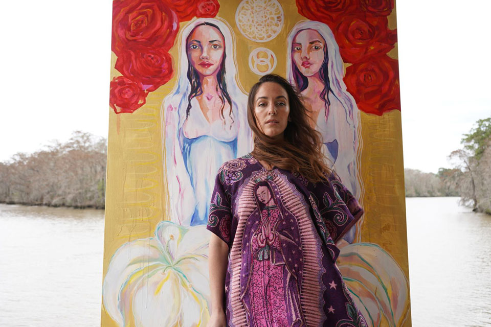 Visionary artist Moon standing in front of her symbolic painting of divine feminine figures, dressed in sacred imagery by the riverside.