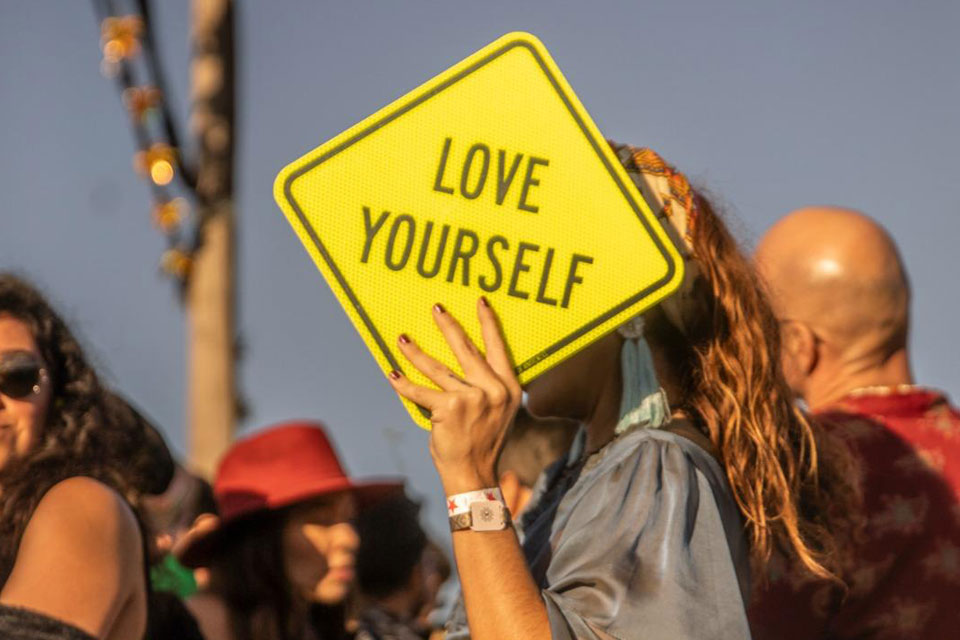 Person holding a neon yellow “LOVE YOURSELF” sign during a public art intervention by Olivia Steele at a cultural gathering.