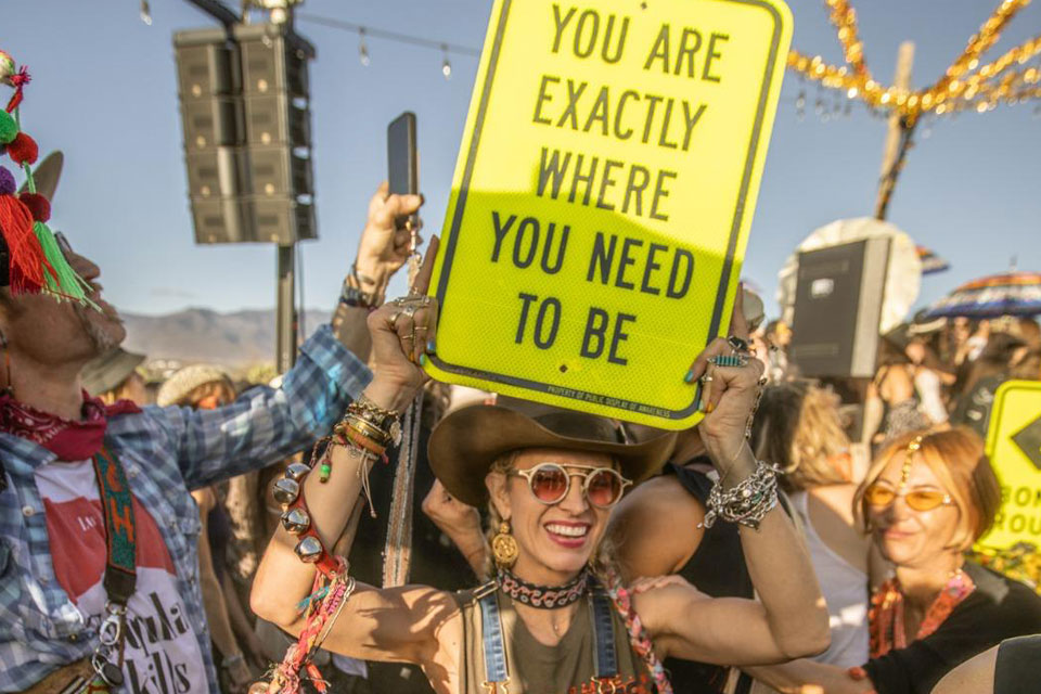 Festival attendee holds up a bright yellow sign that reads "YOU ARE EXACTLY WHERE YOU NEED TO BE" in a joyful crowd.